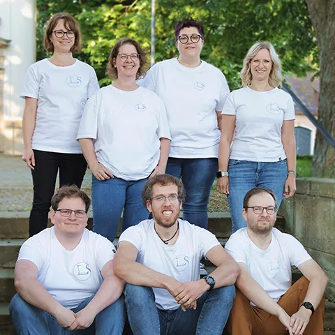 7 Mitarbeiter frontal fotografiert in weißen T-Shirts auf einer Treppe
